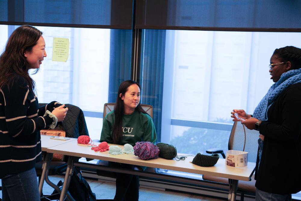 A photo of two people standin in front of a table with knitting supplies and knitted toys on it. One person is sitting behind the table. 
