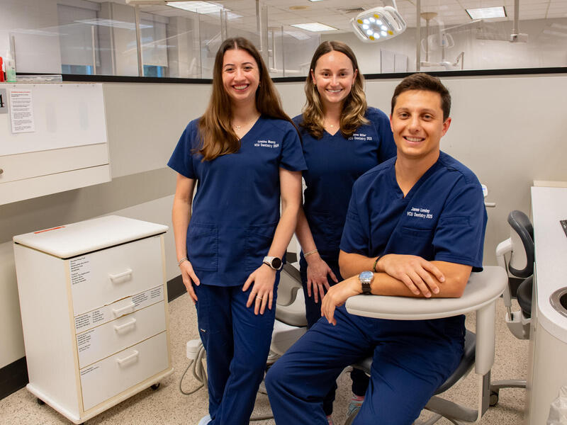 A photo of two women standing and one man sitting in a dental examination room. 