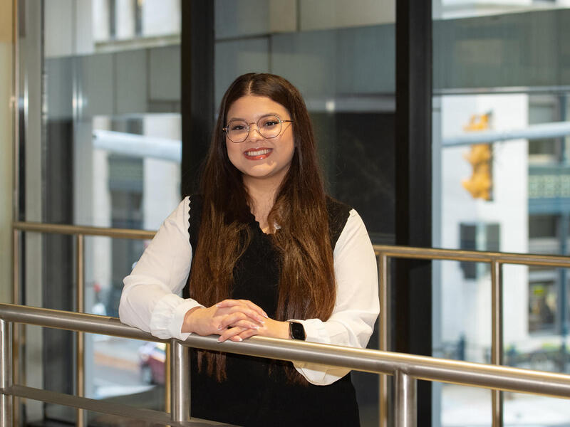 A photo of a woman from the waist up. She is smiling and resting both hands on a metal railing in front of her. 