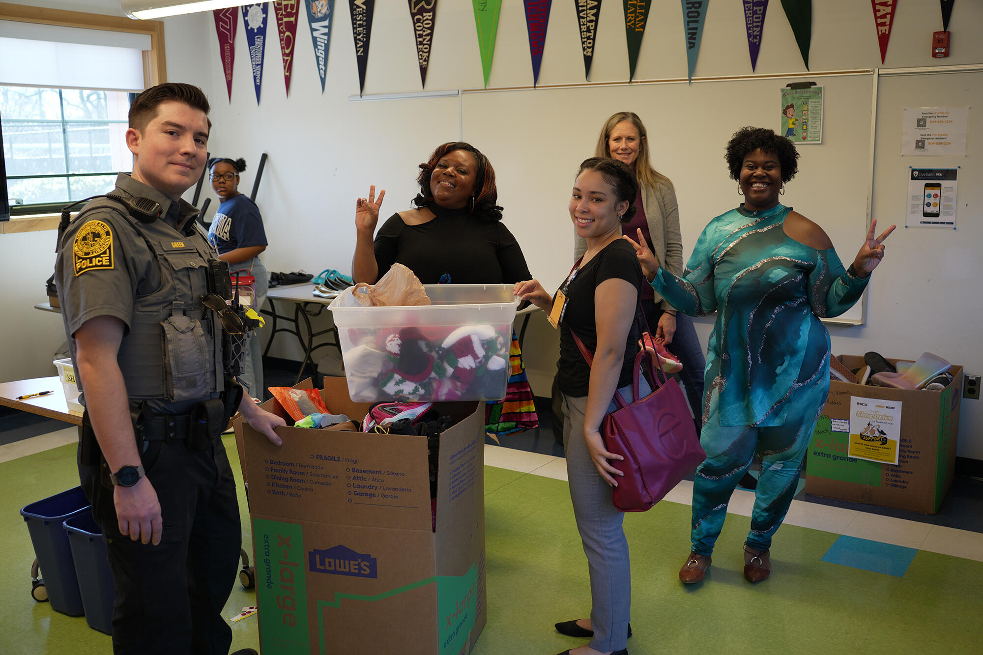 A photo of a police officer standing in a room with five other people. There are three boxes filled with shoes in the room as well. 