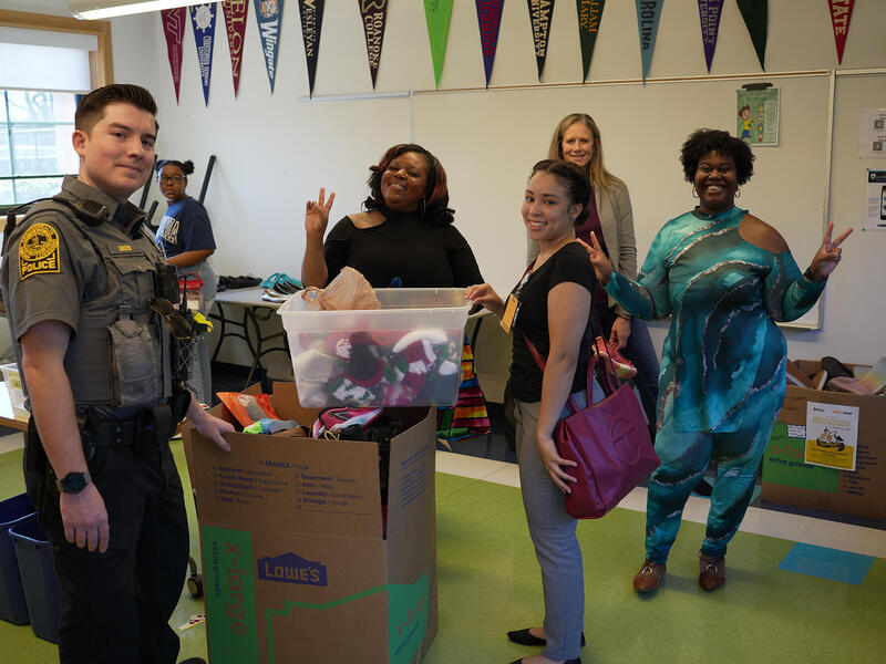 A photo of a police officer standing in a room with five other people. There are three boxes filled with shoes in the room as well. 