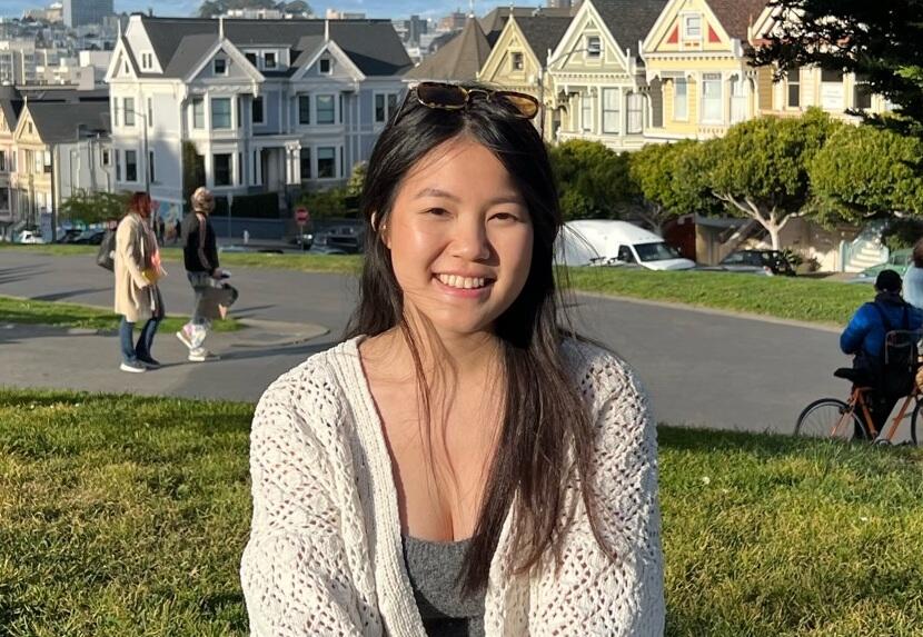 Julie Nguyen sitting on grass with her legs crossed. In the background is a road and rows of homes in a suburban looking area. 