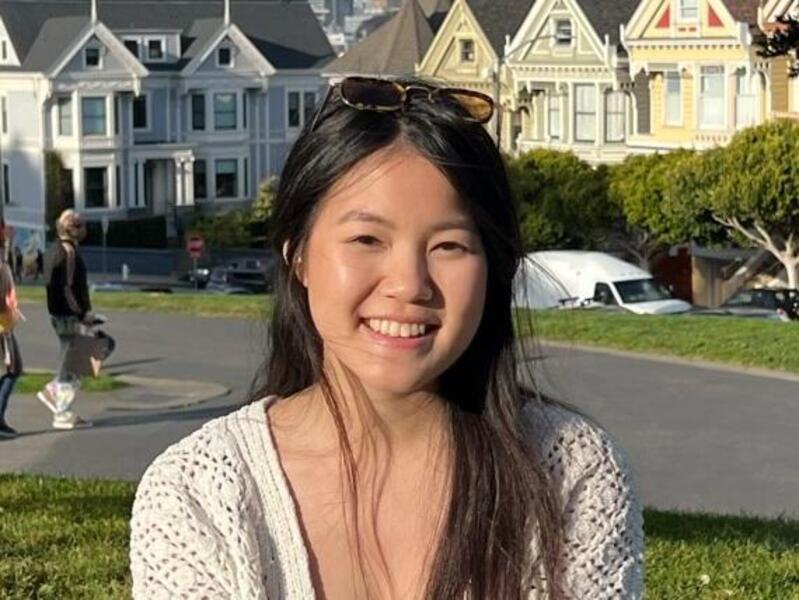Julie Nguyen sitting on grass with her legs crossed. In the background is a road and rows of homes in a suburban looking area. 
