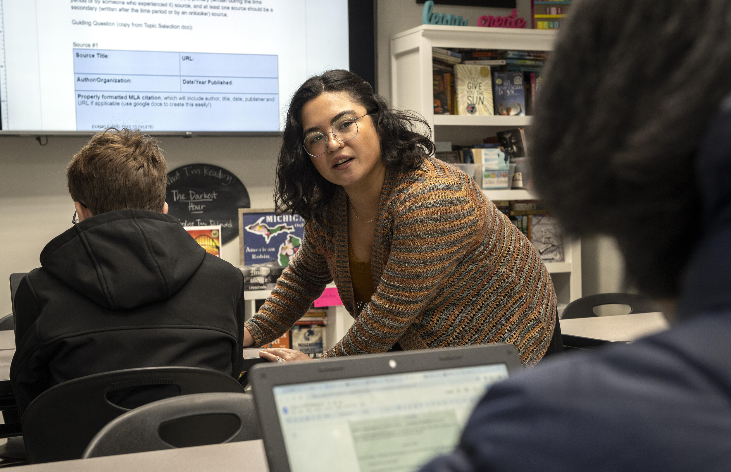 A photo of a woman taking to a student behind her as she leans over next to another sudent's desk. 