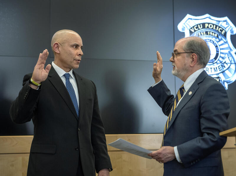 A photo of two men facing each other, both with one arm bent at the elbow with their hand up in the air. The man on the right is holding a stack of papers in his other hand. Both men are wearing suit and ties. 