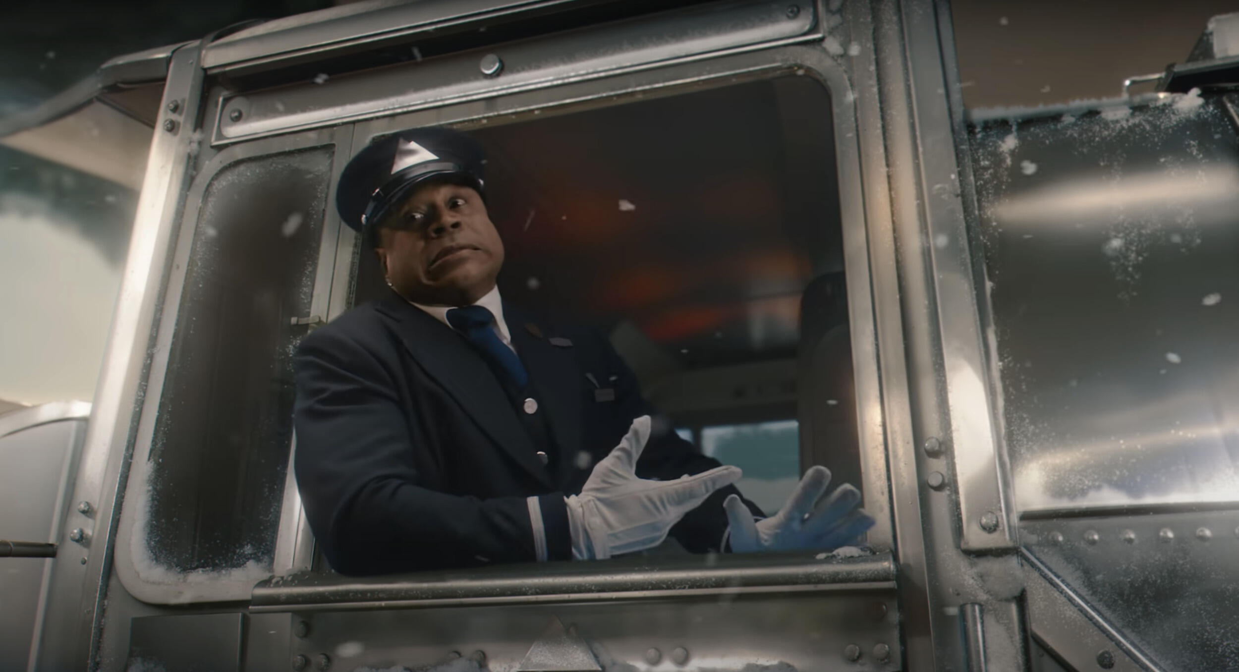 A photo of a man wearing a train conductor univorm sitting in a train engine and looking out the window. 