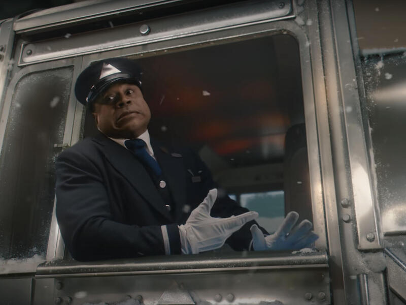 A photo of a man wearing a train conductor univorm sitting in a train engine and looking out the window. 