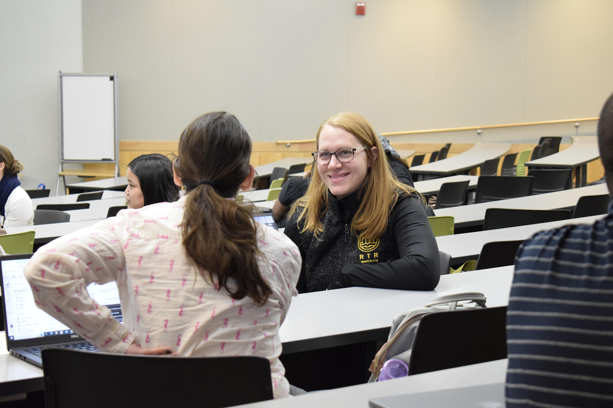 A photo of two people sitting at tables that are next to each other. One person has turned around to face the person sitting behind them. 