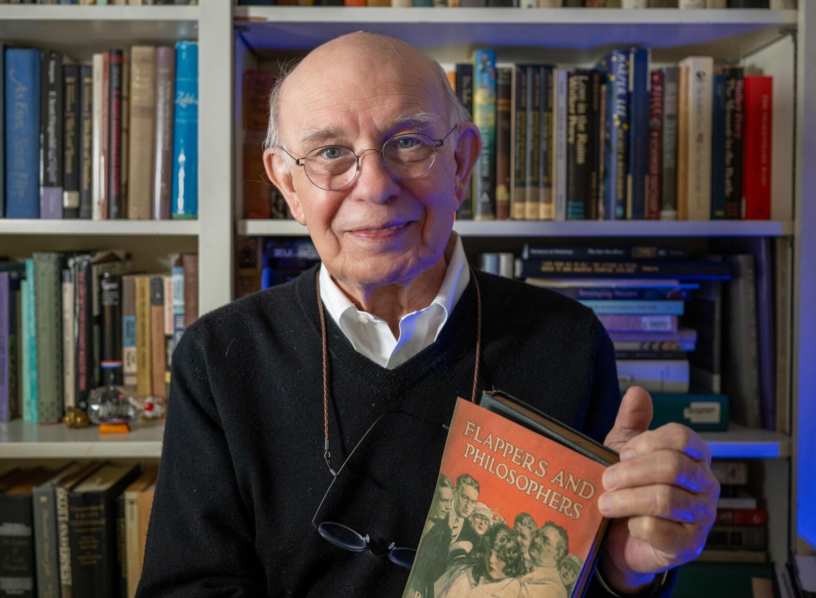 A photo of a man holding a book in front of a book shelf. 