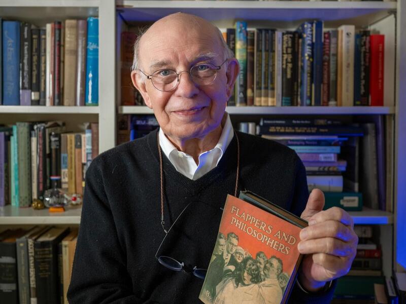 A photo of a man holding a book in front of a book shelf. 