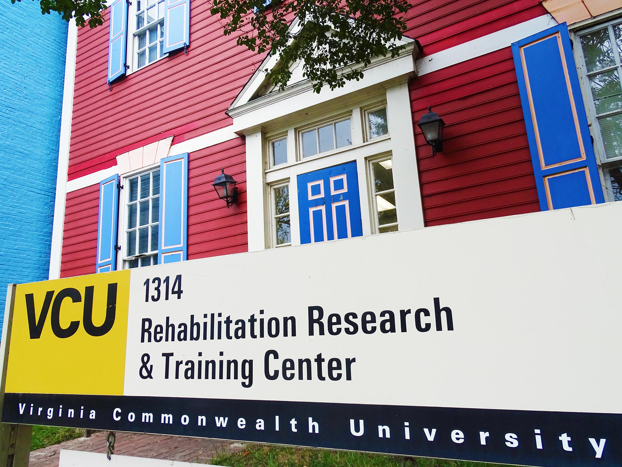 A photo of a sign that says VCU 1314 Rehabilitation Research & Training Center Virginia Commonwealth University. The sign is in front of a red house with blue doors and window shutters.