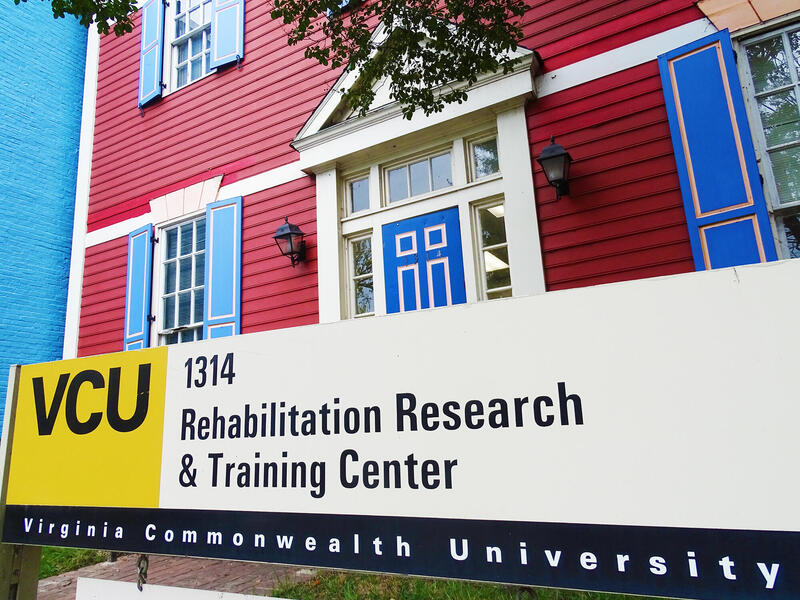 A photo of a sign that says VCU 1314 Rehabilitation Research & Training Center Virginia Commonwealth University. The sign is in front of a red house with blue doors and window shutters.