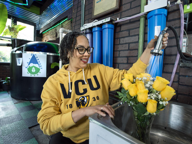 A photo of a woman using a sink hose to fill a vase full of yellow roses that are sitting in a sink. 