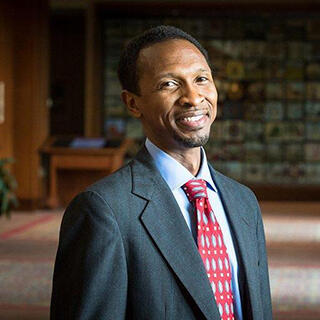 A portrait of a man wearing a black sports coat, light blue button up shirt, and a dark red tie with a white and light blue pattern.;