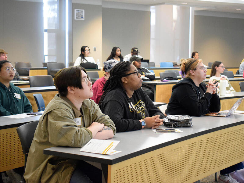 A photo of rows of desks and chairs. People are spread out throughout the rows sitting at the tables. 