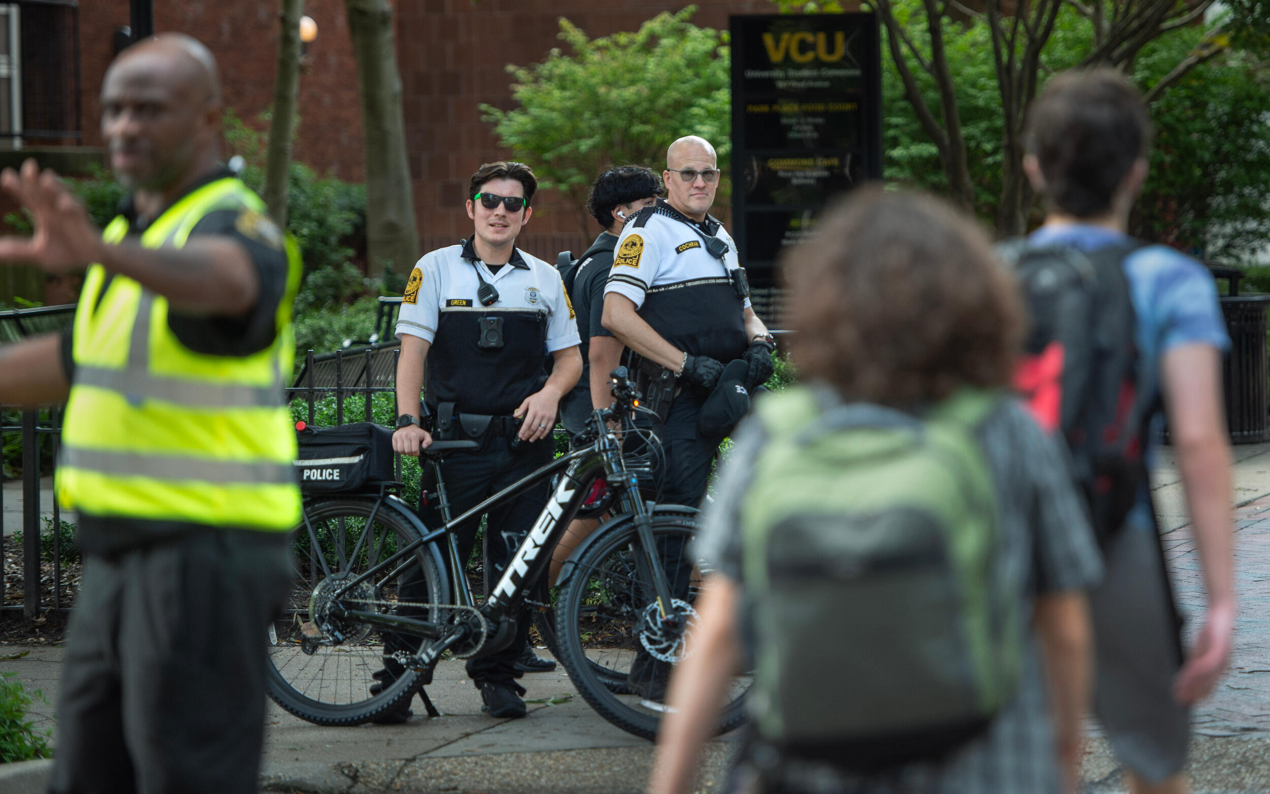 Two police officers with bicycles look toward the camera while students walk around them.