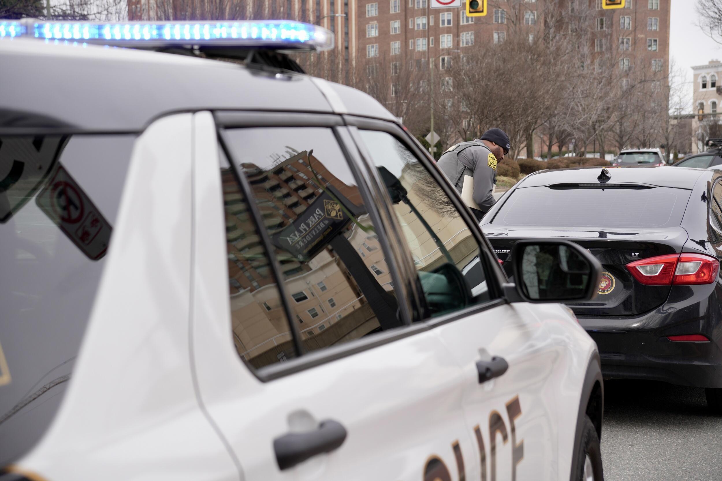A photo of a police car parked behind a black sedan. A police officer is standing on the driver side of the black car. 