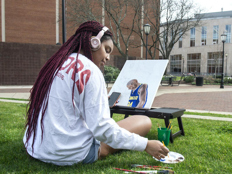 A student sitting in the grass on a warm day while painting outside in Park Plaza.
