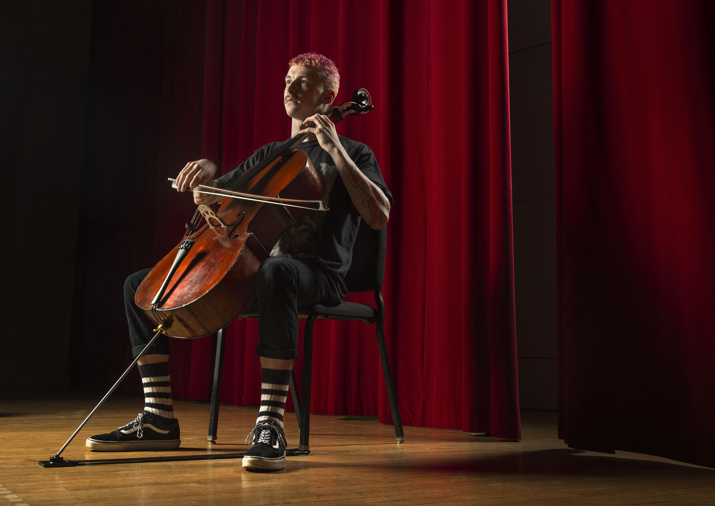 A man sitting in a chair in front of a red curtain playing the chelo. 