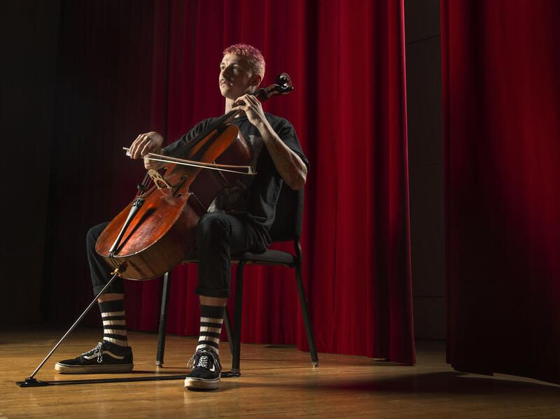 A man sitting in a chair in front of a red curtain playing the chelo. 
