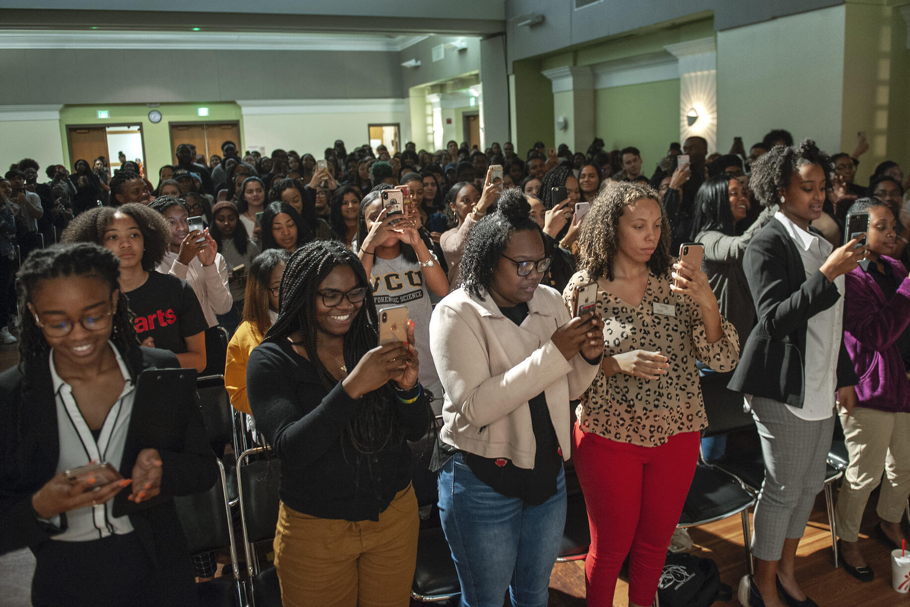 Attendees of a speaking event stand. Some attendees are taking photos on cell phones.