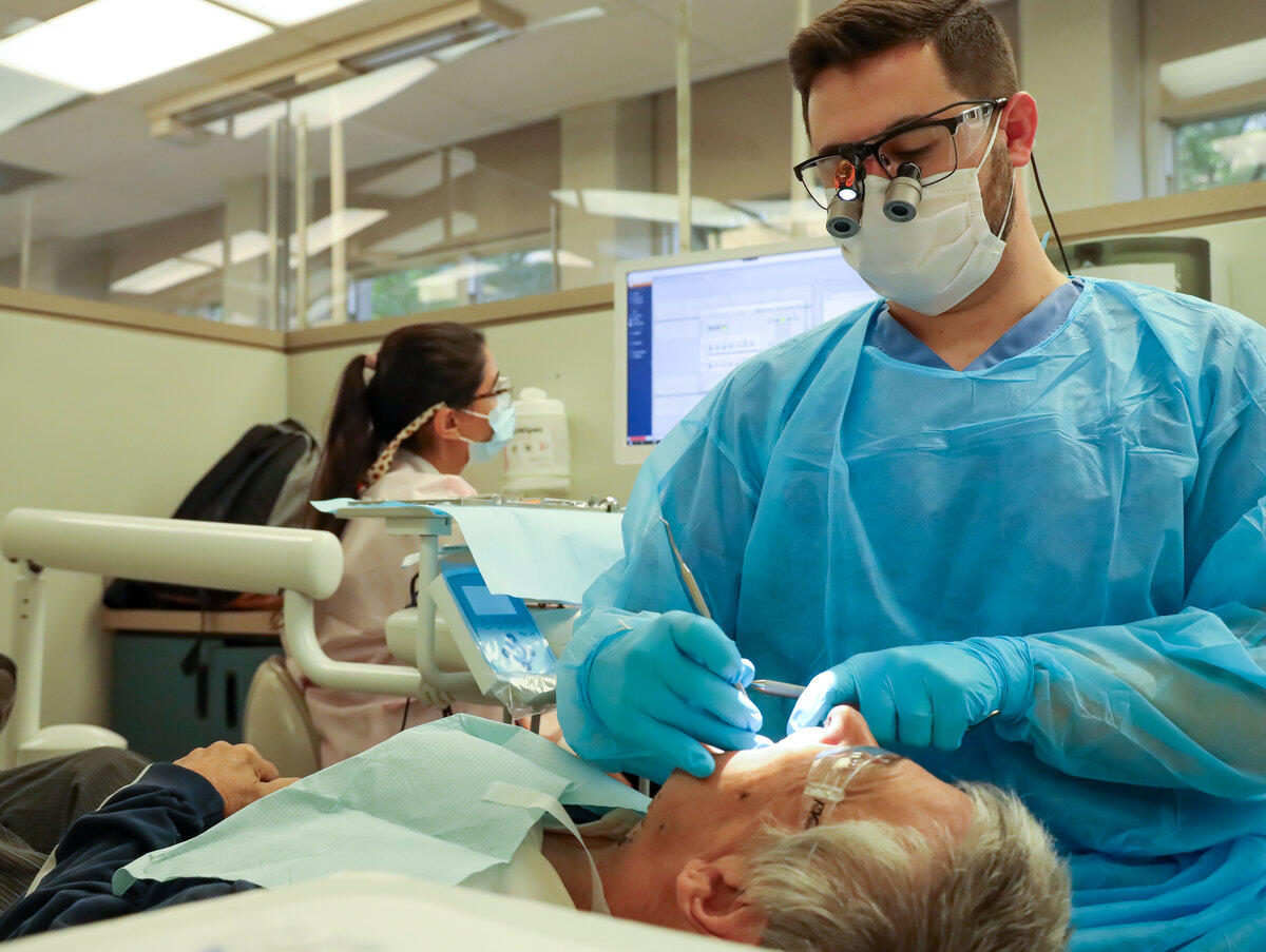 A photo of a man in blue scrubs cleaning the teeth of a man who is laying down in front of him. 