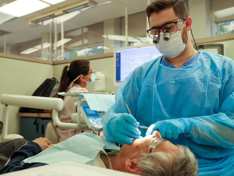 A photo of a man in blue scrubs cleaning the teeth of a man who is laying down in front of him. 