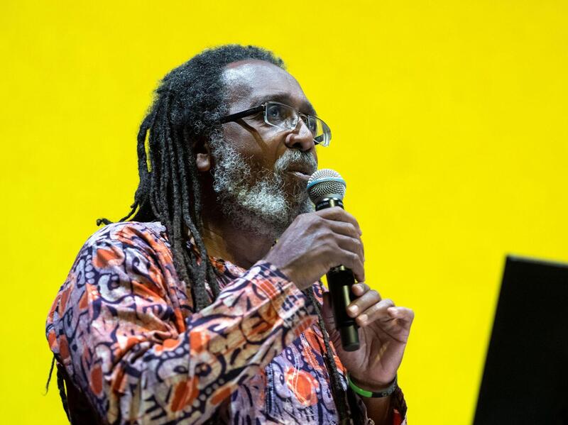 A man with long dreads speaking into a microphone in front of a yellow background. 
