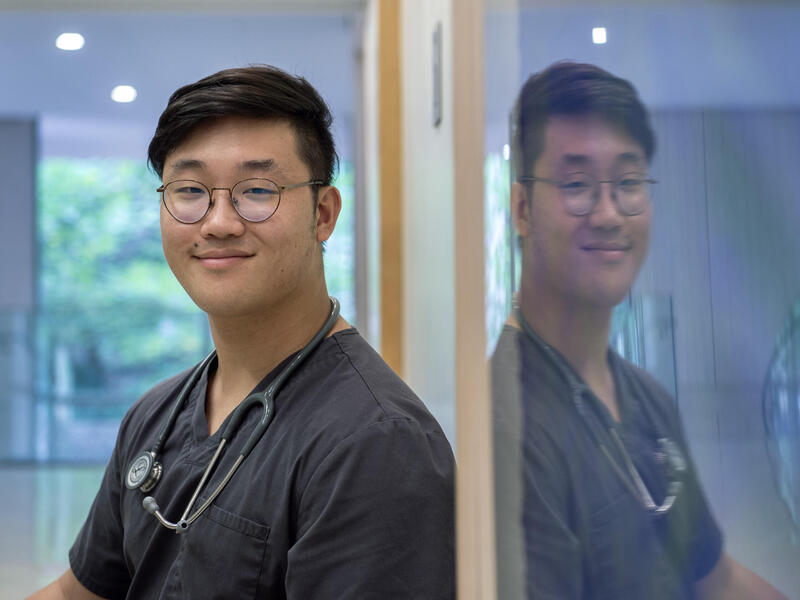 A photo of a man from the chest up standing next to a wall with glass on it. The glass is reflecting the image of the man on it. The man is wearing a medical scrub shirt and a stethoscope around his neck. 
