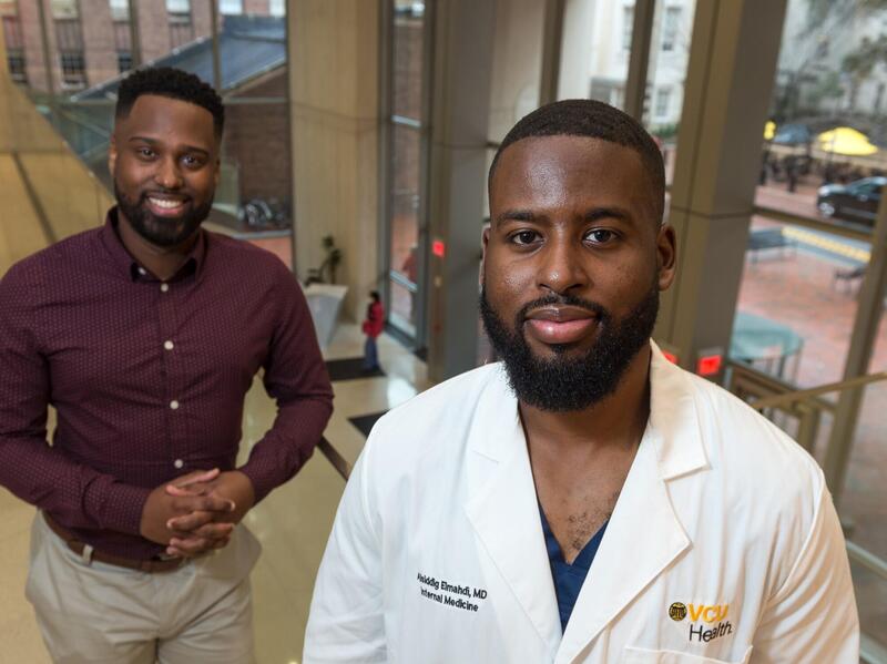 Two men, one in a doctor's coat, pose for a photo in a hallway in front of windows.