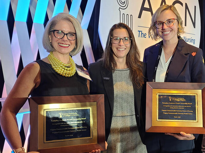 A photo of three women from the waist up. The women on the far left and right are holding award plaques. 
