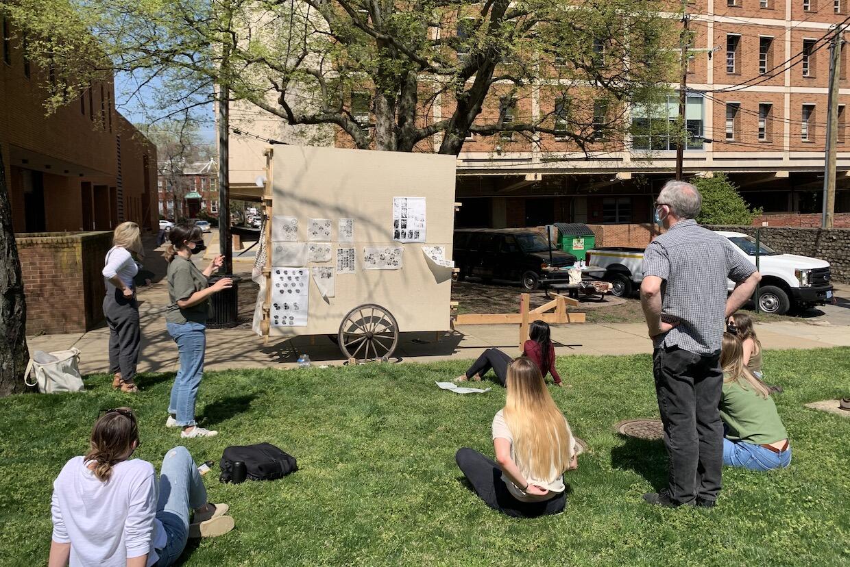 Emily Smith (standing left in jeans) and Camden Whitehead (standing on the right in jeans) lead an interior design department class critique outside using a mobile cart where they can pin up student work.