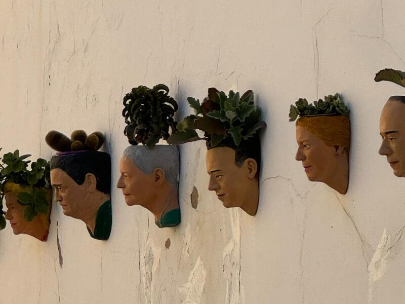 A photo of a wall with planters hanging on it. The planters are shaped like human faces and have a plant sticking out of the top. 