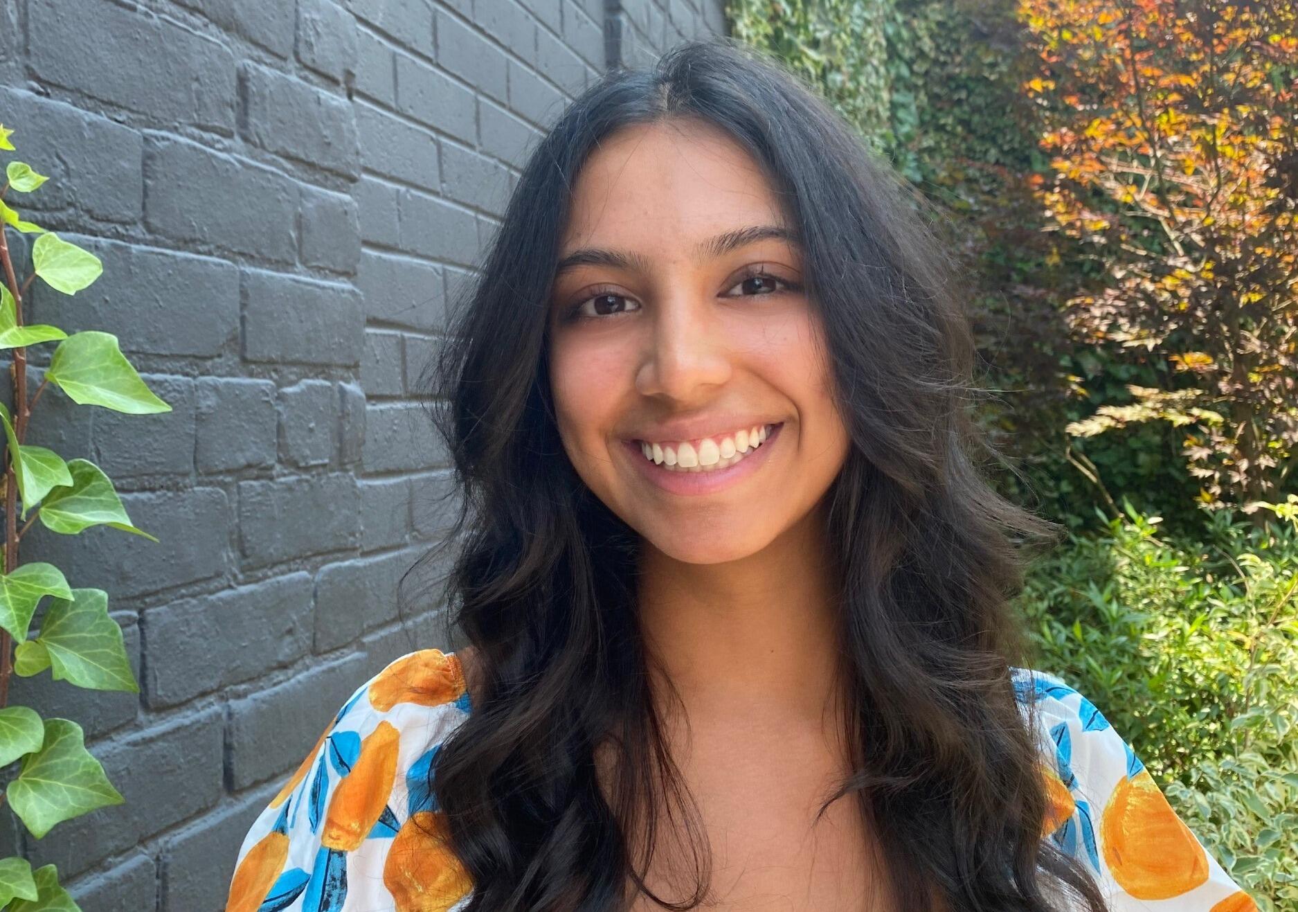 A portrait of Avani Venkatesh standing next to a black brick wall with vines growing up the side of it 