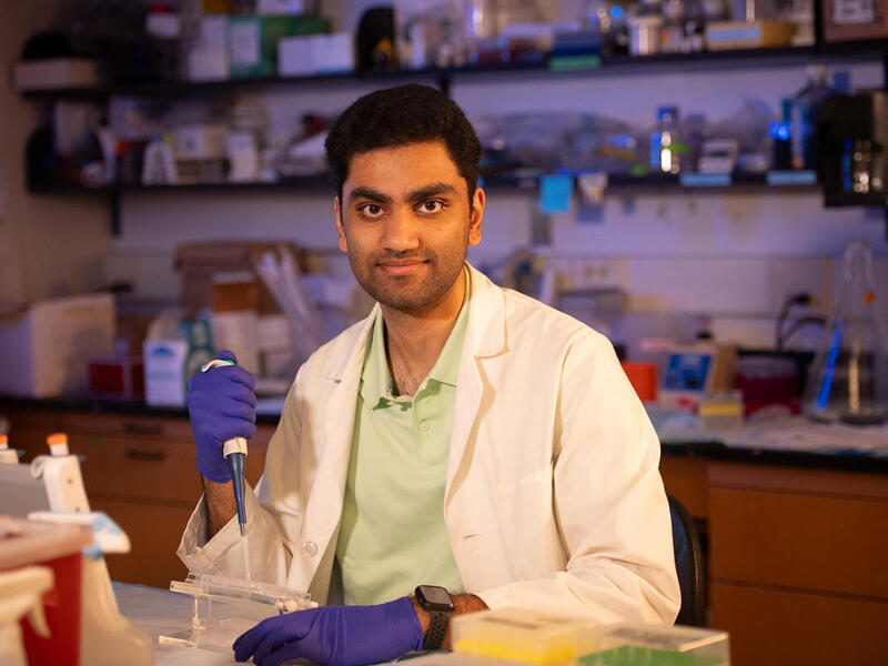 A photo of a man wearing a white lab coat and blue gloves sitting at a table. In his  left hand he is holding a dropper. 