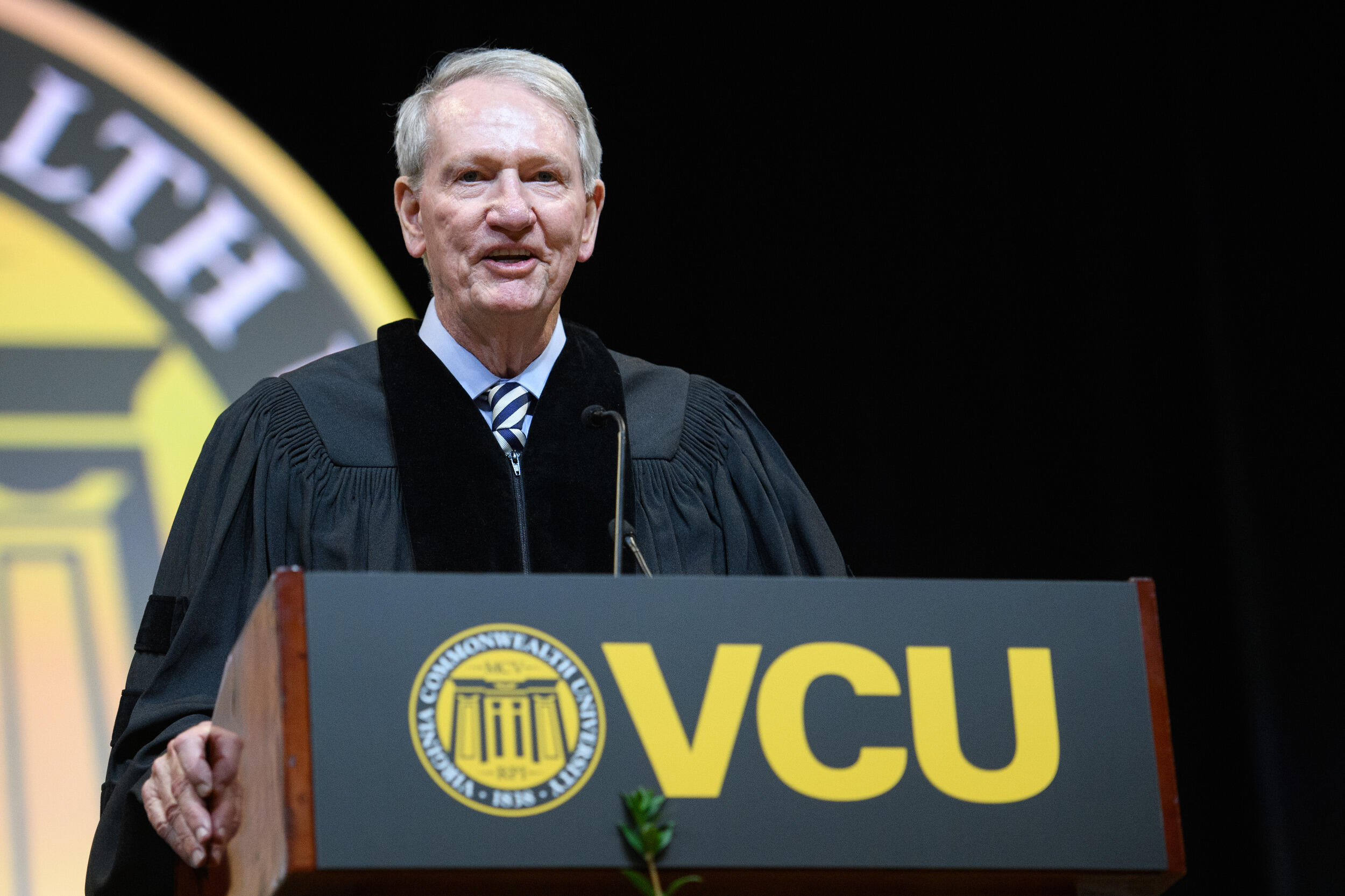 A man in a graduation gown speaks at a lectern with a VCU sign and logo on the front of it.