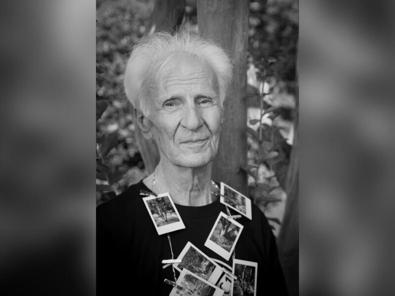 A black and white photo of an old man from the chest up. Photographs are pinned to his black shirt 