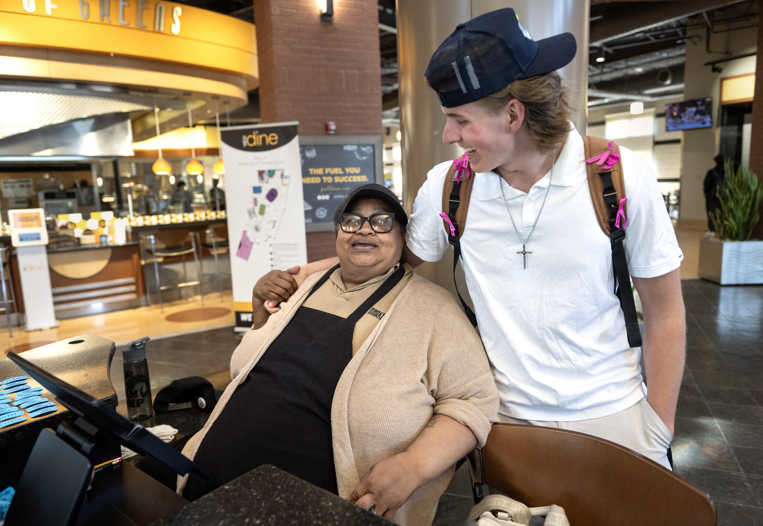 A photo of a woman sitting. Next to her is a young man with his arm around her shoulder. 