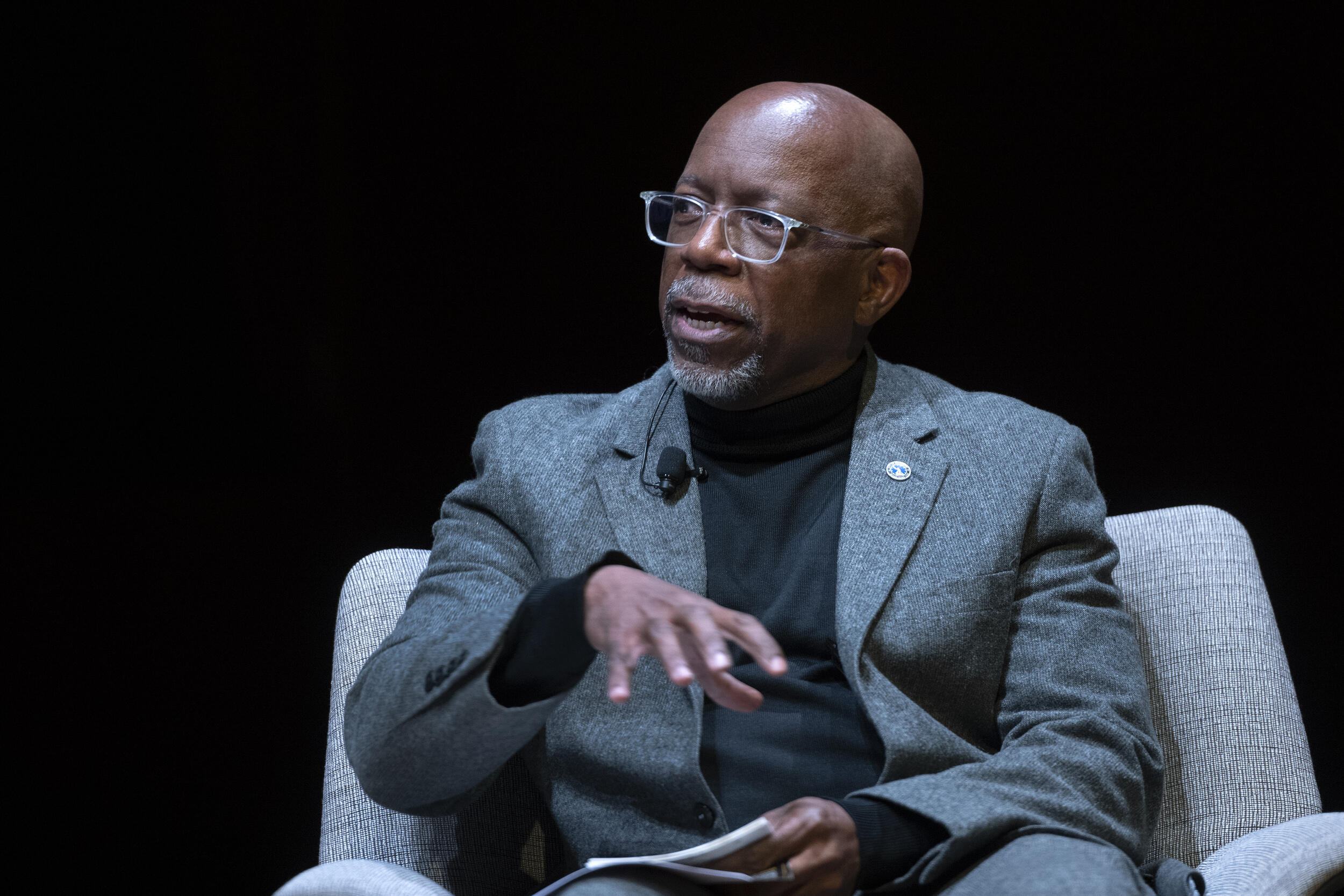 A bald man wearing a gray sportscoat, black turtleneck shirt and classes sitting in a chair looking to his right. 