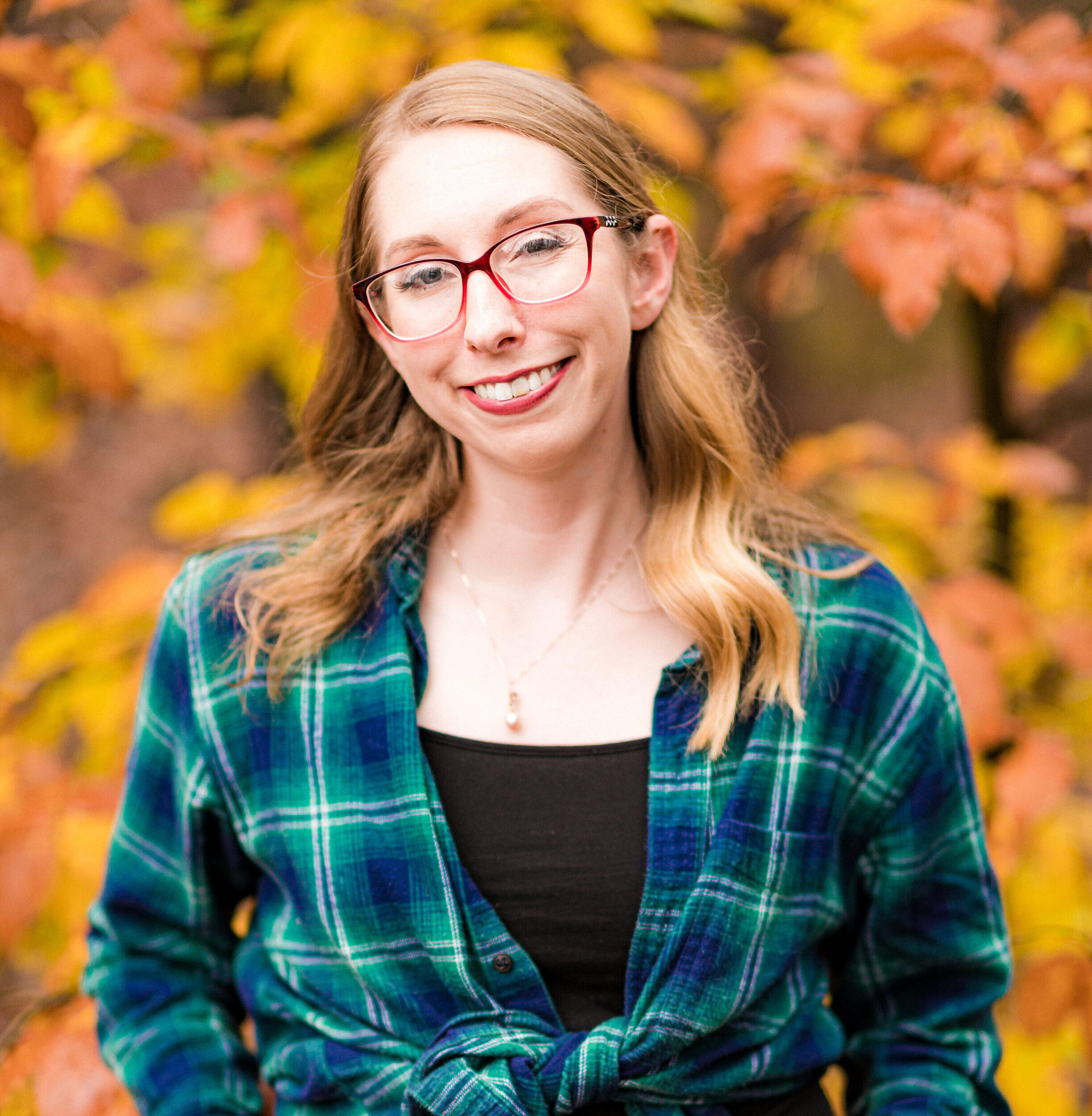 A woman in glasses and a flannel shirt smiles with fall leaves behind her.