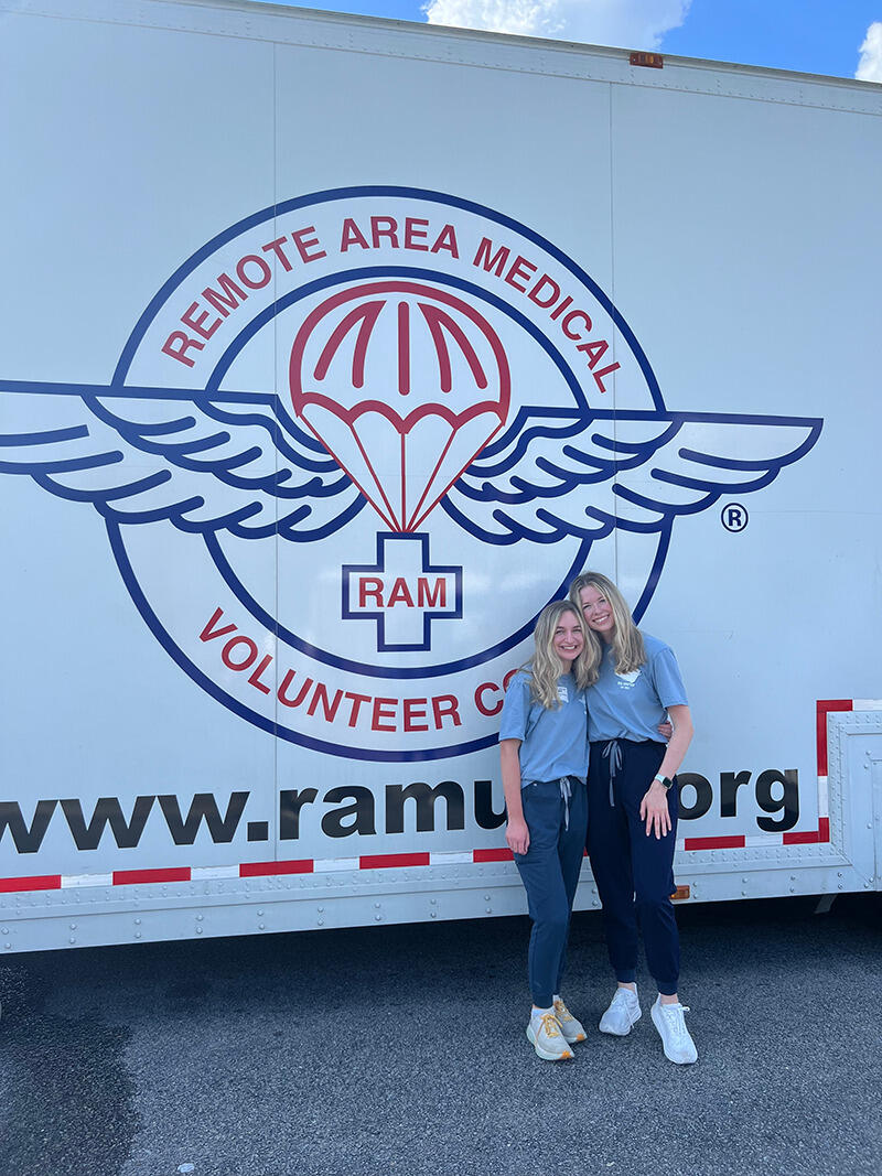 A photo of two women standing in front of a trailer that says \"REMOTE AREA MEDICAL VOLUNTEER\" 