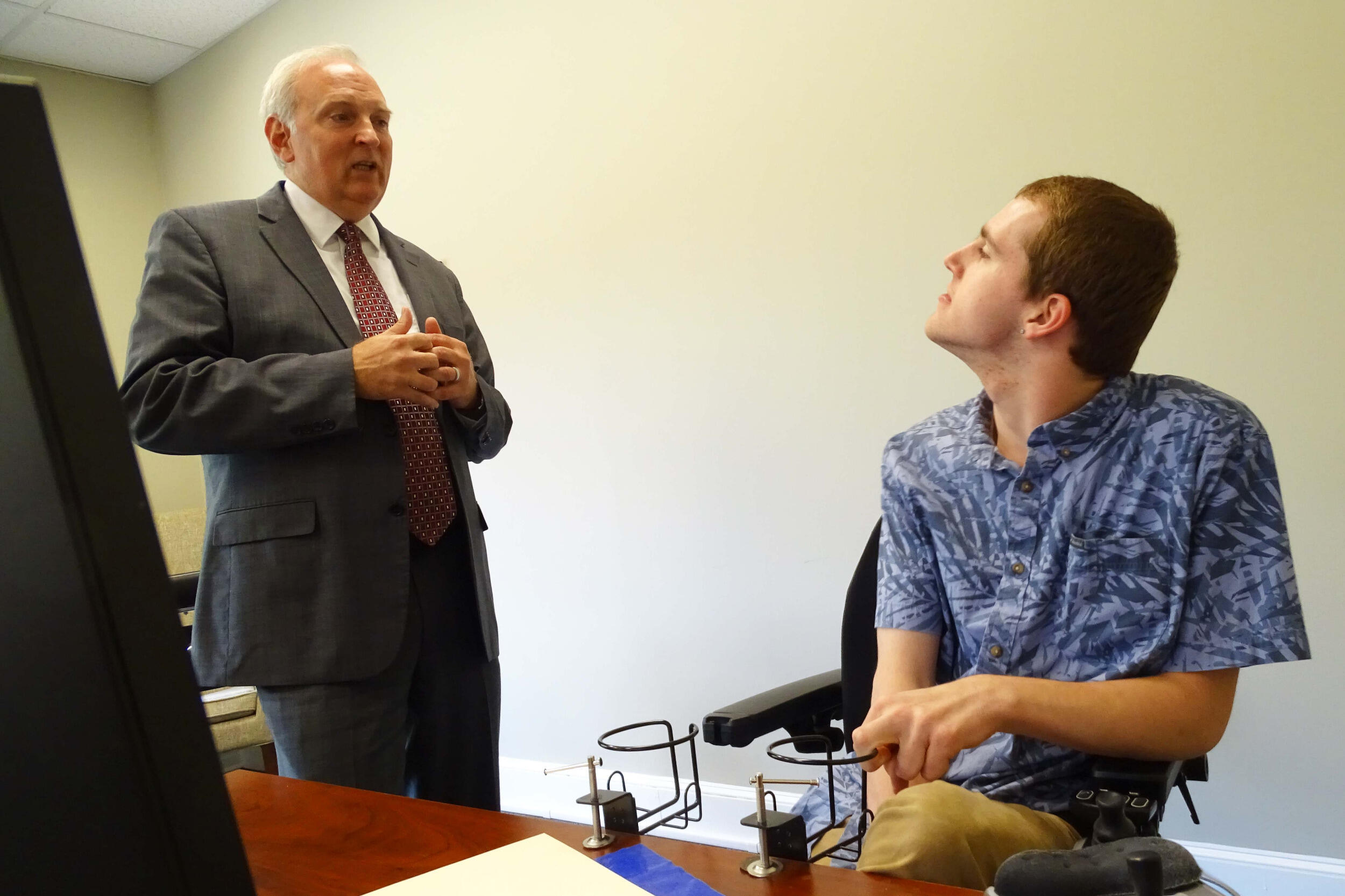 A man in a coat and tie stands and speaks to a man who is sitting in front of a computer screen.