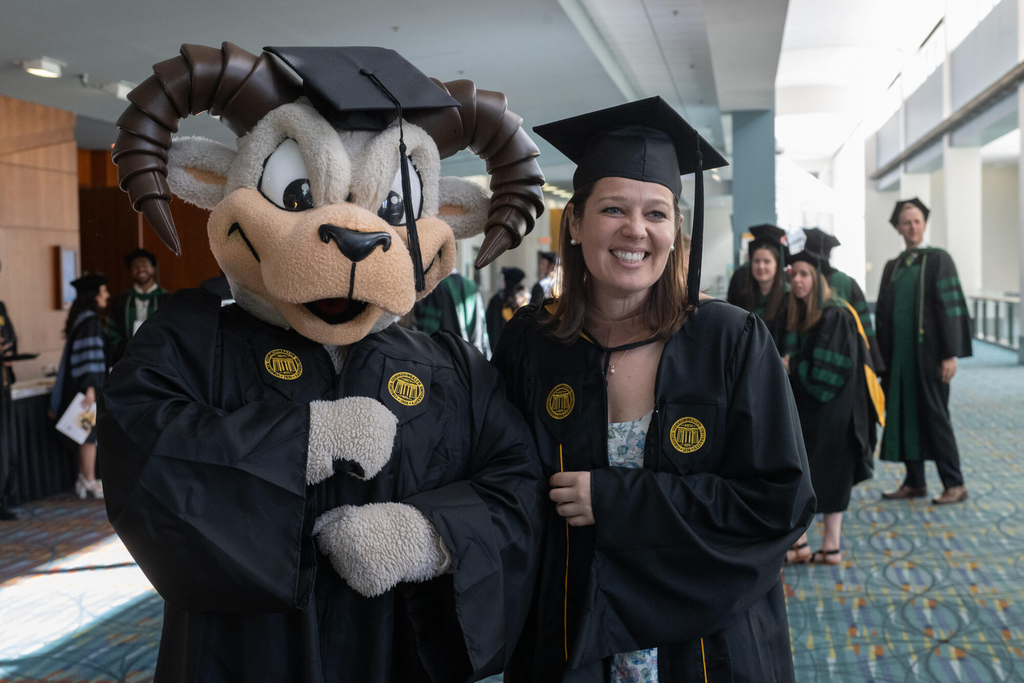 A graduate and mascot Rodney the Ram pose for a picture, both wearing graduation caps and gowns.