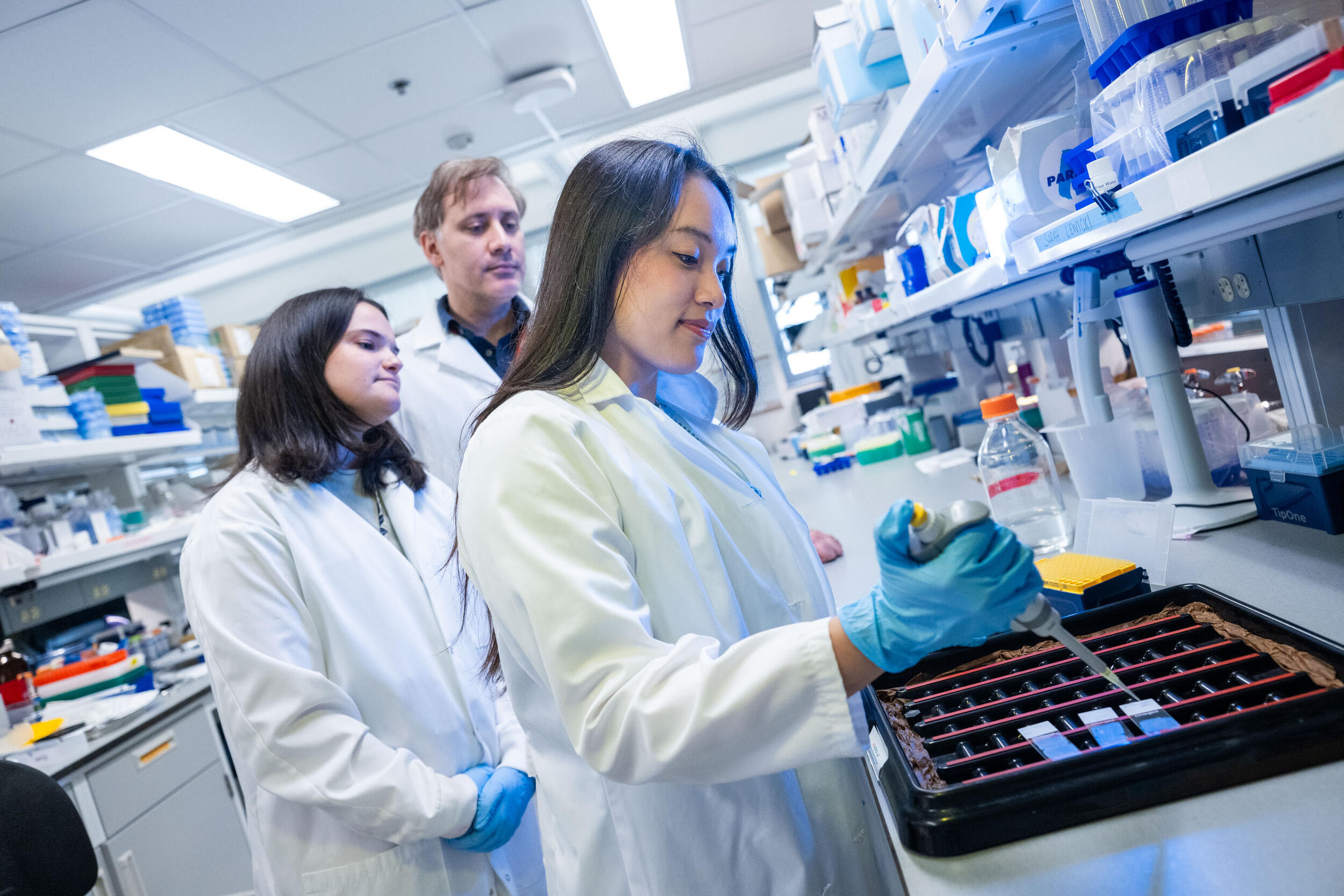 A woman in a lab coat works with a pipette in a lab while a woman in a lab coat and a man in a lab coat watch.