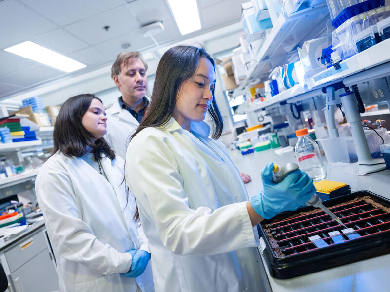 A woman in a lab coat works with a pipette in a lab while a woman in a lab coat and a man in a lab coat watch.
