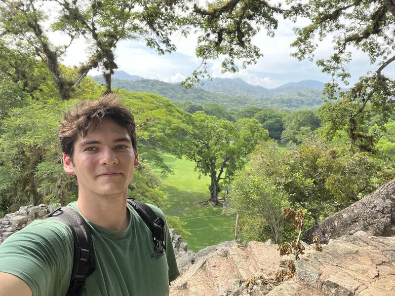 A photo of a man from the chest up standing on a cliff side. Behind him is a grassy area with trees, and further in the distance are mountains 