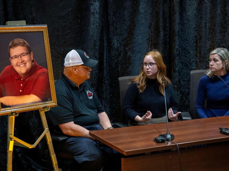 Three people sit at a desk speaking together with a microphone in front of them. To their right is a portrait on an easel of a young man in glasses and a red shirt.