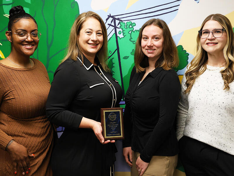 A photo of a four women from the waist up. The second woman to the left is holding a small plaque. 