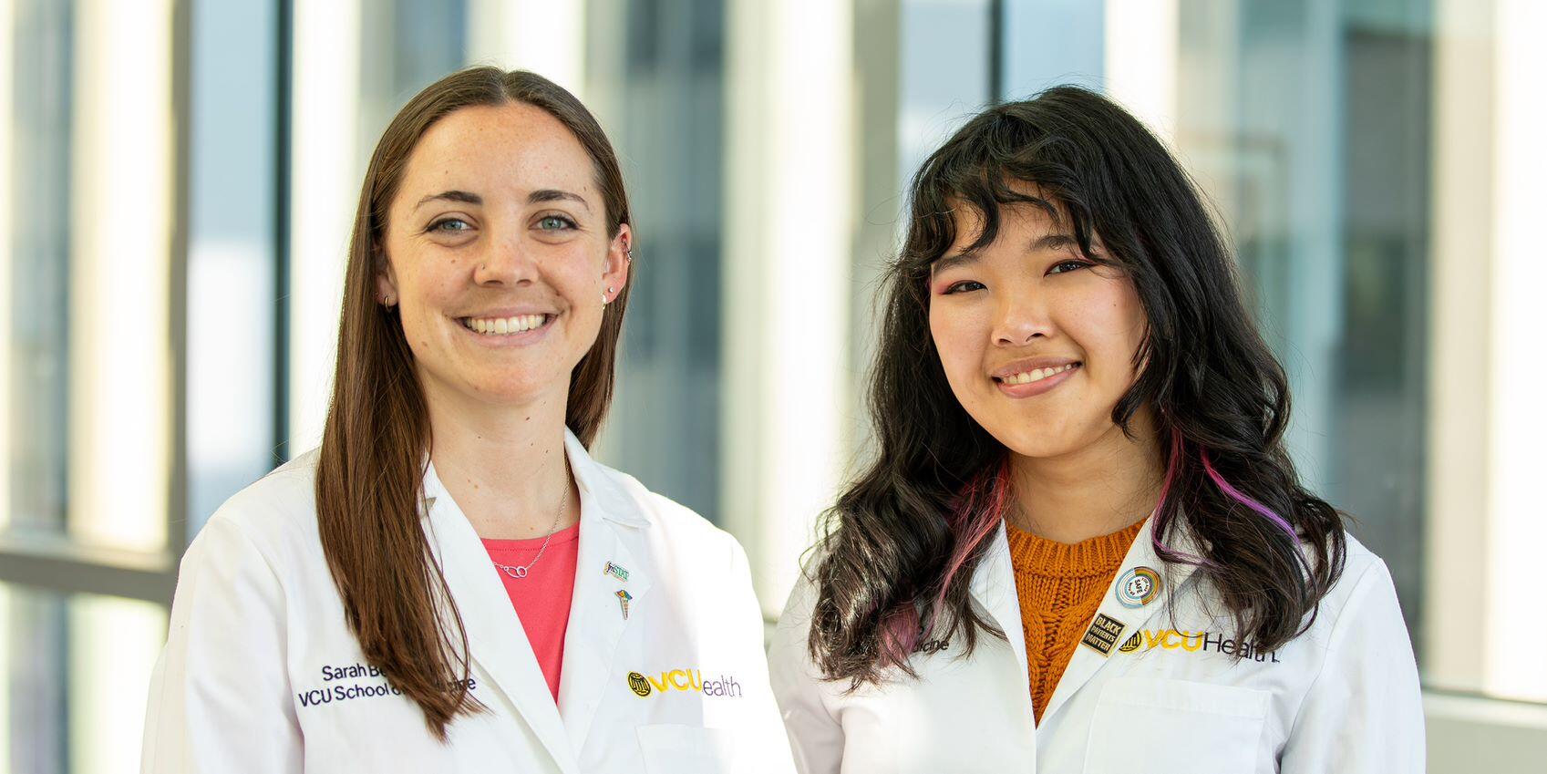 Two women wearing white lab coats standing in front of windows. 