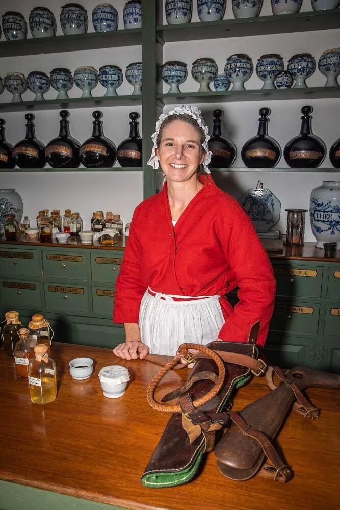 A person wearing colonial-era clothing inside of an apothecary shop. Bottles and wares line the wall behind them and on the table in front.